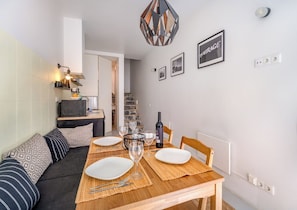 A dining area with a wooden table set for two, a sofa with patterned cushions, a TV, and a staircase leading to another room.