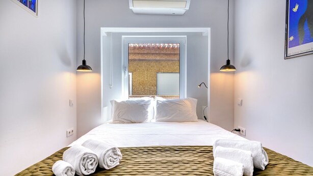 A hotel room with a bed, two bedside lamps, a window with a view, and a wall-mounted air conditioner. - Cozy 1-bedroom house in the historic old town of Portimão. (Portimão)