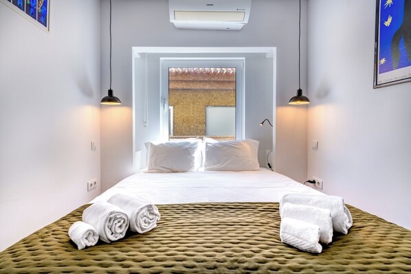 A hotel room with a bed, two bedside lamps, a window with a view, and a wall-mounted air conditioner.