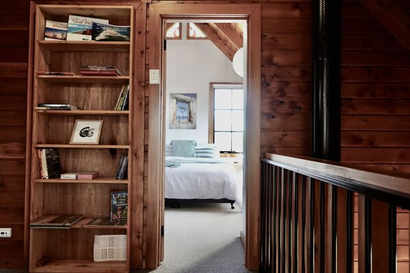 A wooden bookshelf with books and a framed picture, leading to a bedroom with a bed and a window.