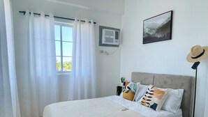 A bedroom with a bed, a window with curtains, a ceiling fan, a wall-mounted air conditioner, a framed picture, and a floor lamp.