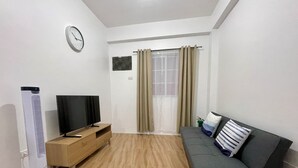A living room with a flat-screen TV on a wooden stand, a sofa with cushions, a wall clock, and a window with curtains.