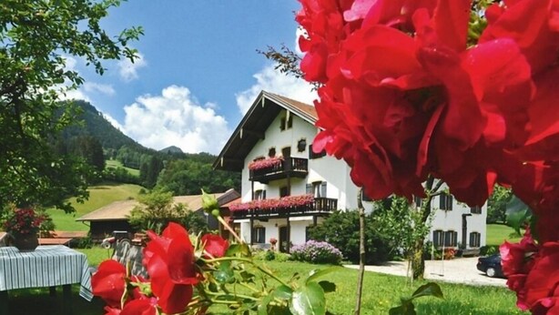 Un bâtiment à deux étages doté d’un balcon, entouré d’une végétation luxuriante et avec des montagnes en arrière-plan.