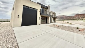 A modern two-story house with a large garage, a balcony, and a view of a desert landscape. - Luxury Kanab Retreat | Pool, Hot Tub Near Zion (Kanab)