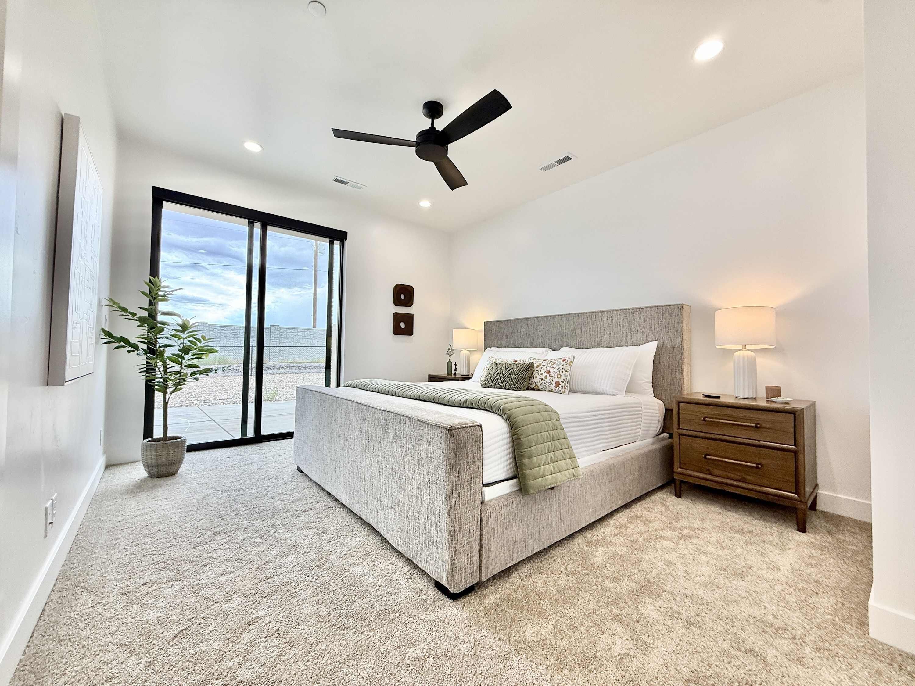 A bedroom with a bed, a nightstand, a lamp, a ceiling fan, and a large window with a view of the outdoors.