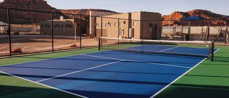Una cancha de tenis al aire libre con una red, rodeada por una valla y edificios al fondo.