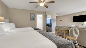 A hotel room with two beds, a desk, a chair, a ceiling fan, a TV, and a balcony with a view.
