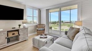 A living room with a TV, a sofa, a chair, a small table, and a balcony with a view.