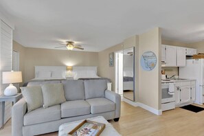 Ein modernes, gut beleuchtetes Wohnzimmer mit einer Couch, einem Deckenventilator und einer Küchenzeile mit weißen Schränken und einem Kühlschrank.