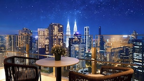 Balcón alto con sillas de mimbre y una pequeña mesa, con vista a un paisaje urbano nocturno.