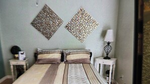 Una camera da letto con un letto, un ventilatore a soffitto, due elementi decorativi a parete, un comodino con una lampada e un tavolino di sostegno.