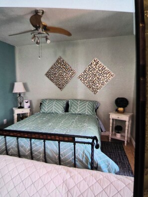 A bedroom with a bed, a ceiling fan, two decorative wall pieces, a nightstand with a lamp, and a small table with a plant.