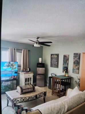 A living room with a sofa, a coffee table, a TV, a ceiling fan, a refrigerator, and a dining table with chairs.