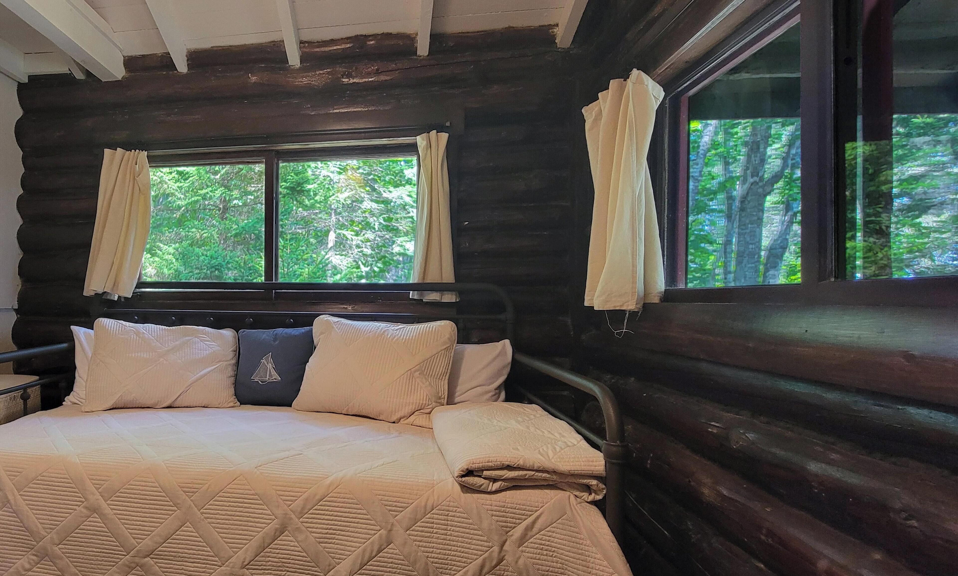 A wooden cabin bedroom with a bed, curtains, and a window overlooking greenery.