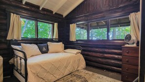 A log cabin bedroom with a bed, two windows with curtains, a dresser, and a fan. - Escape to The Hideout, a charming 2-bedroom cabin in Boothbay Harbor. (Boothbay Harbor)