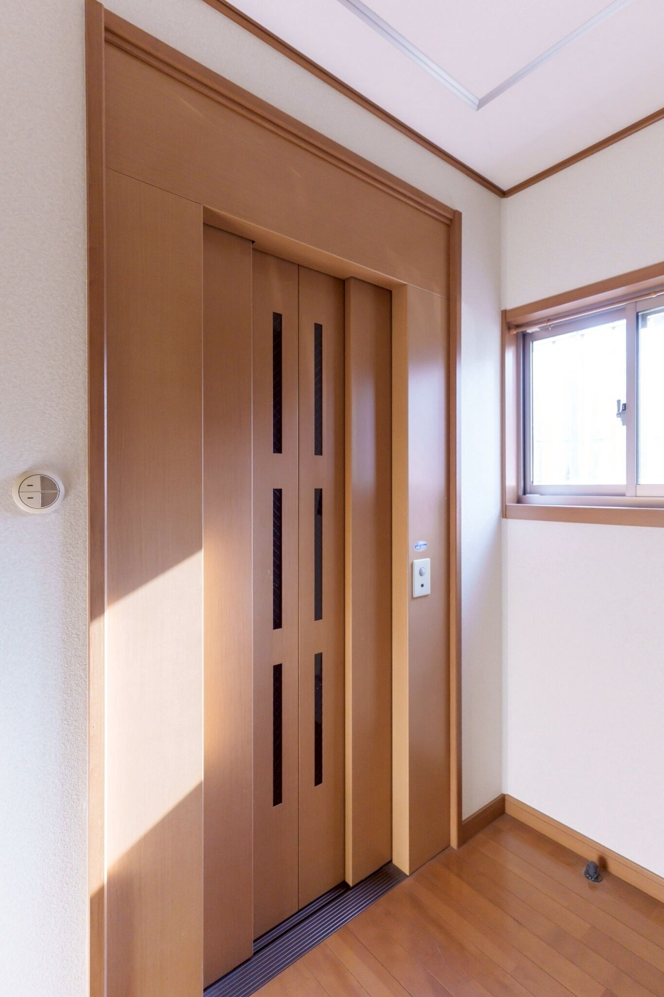 A wooden elevator door with a vent, a window to the right, and a light switch on the wall.