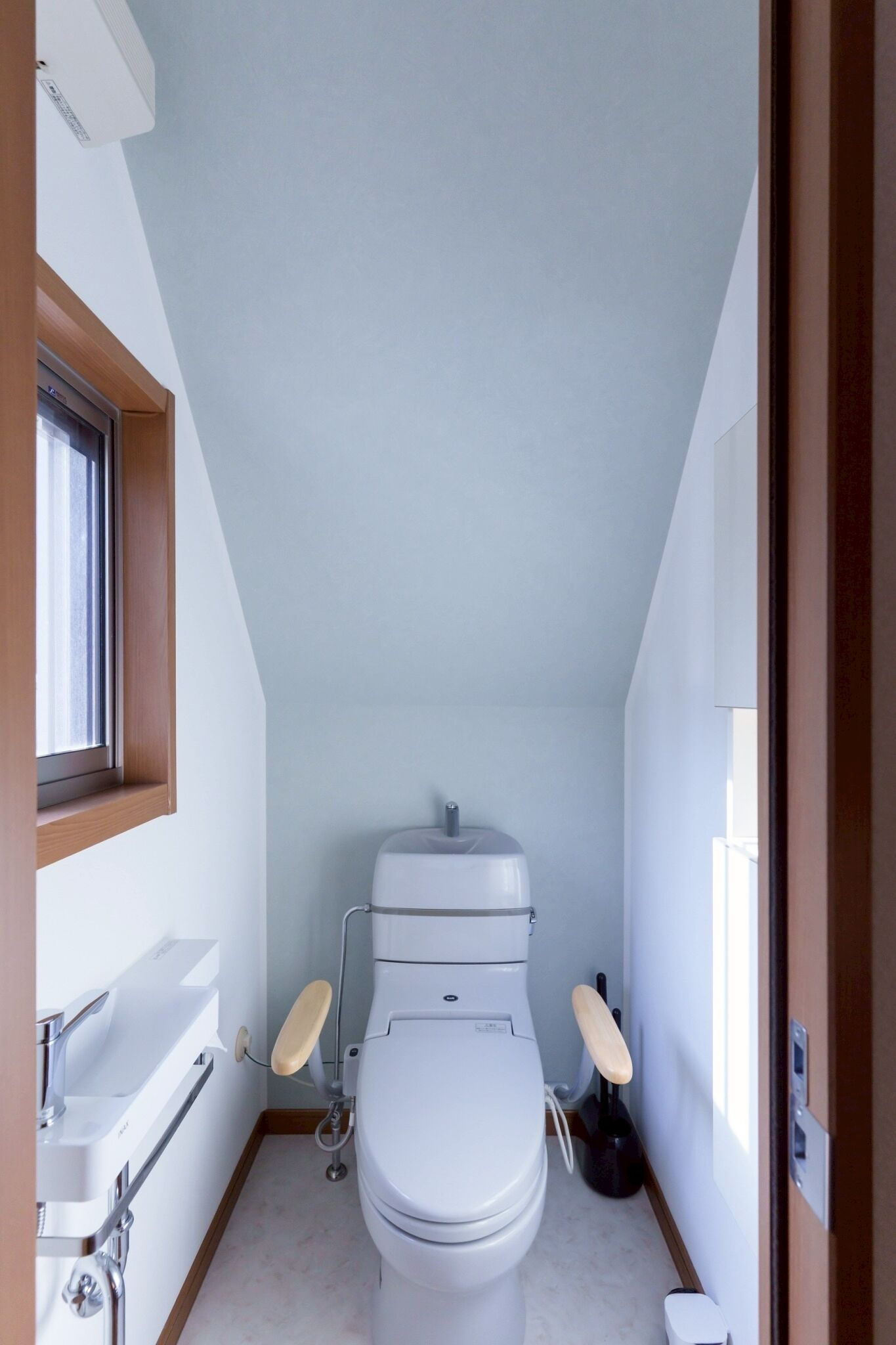 A small, narrow bathroom with a white toilet, a wooden-framed window, and a white sink.