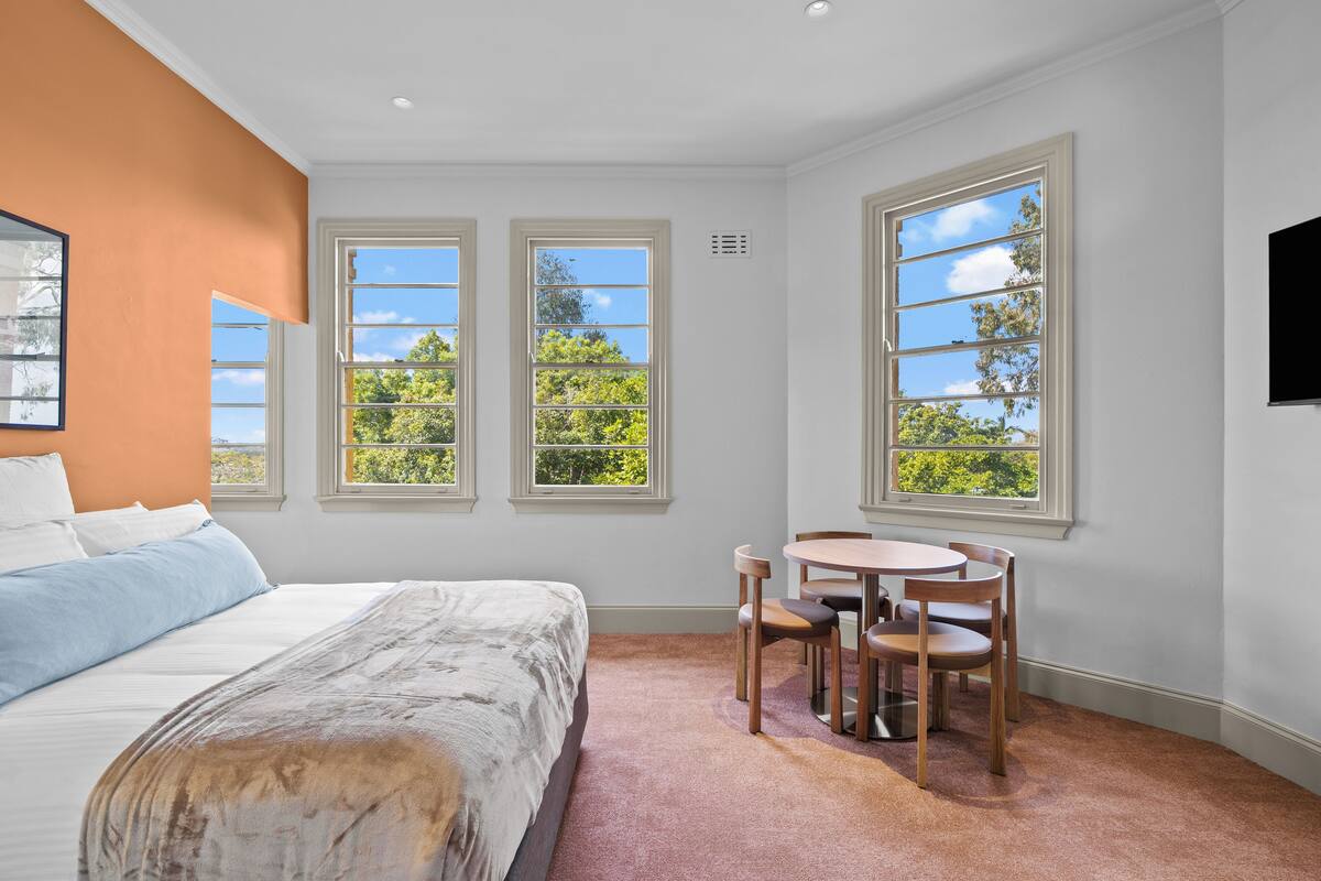 A bedroom with a bed, a desk with chairs, and large windows with a view of trees.