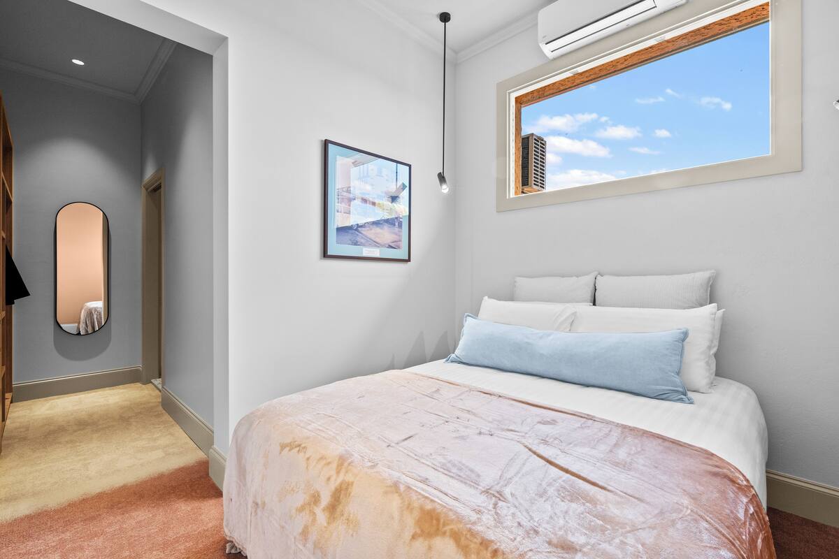 A bedroom with a bed, a framed picture, a window with a view, and a mirror.