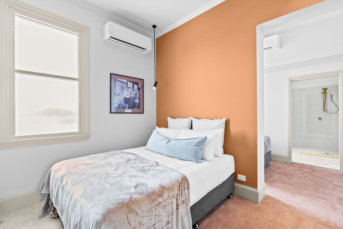 A bedroom with a bed, an air conditioning unit, a framed picture on the wall, and a door leading to a bathroom.