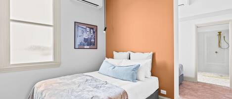 Un dormitorio con cama, unidad de aire acondicionado, un cuadro enmarcado en la pared y una puerta que da al baño.