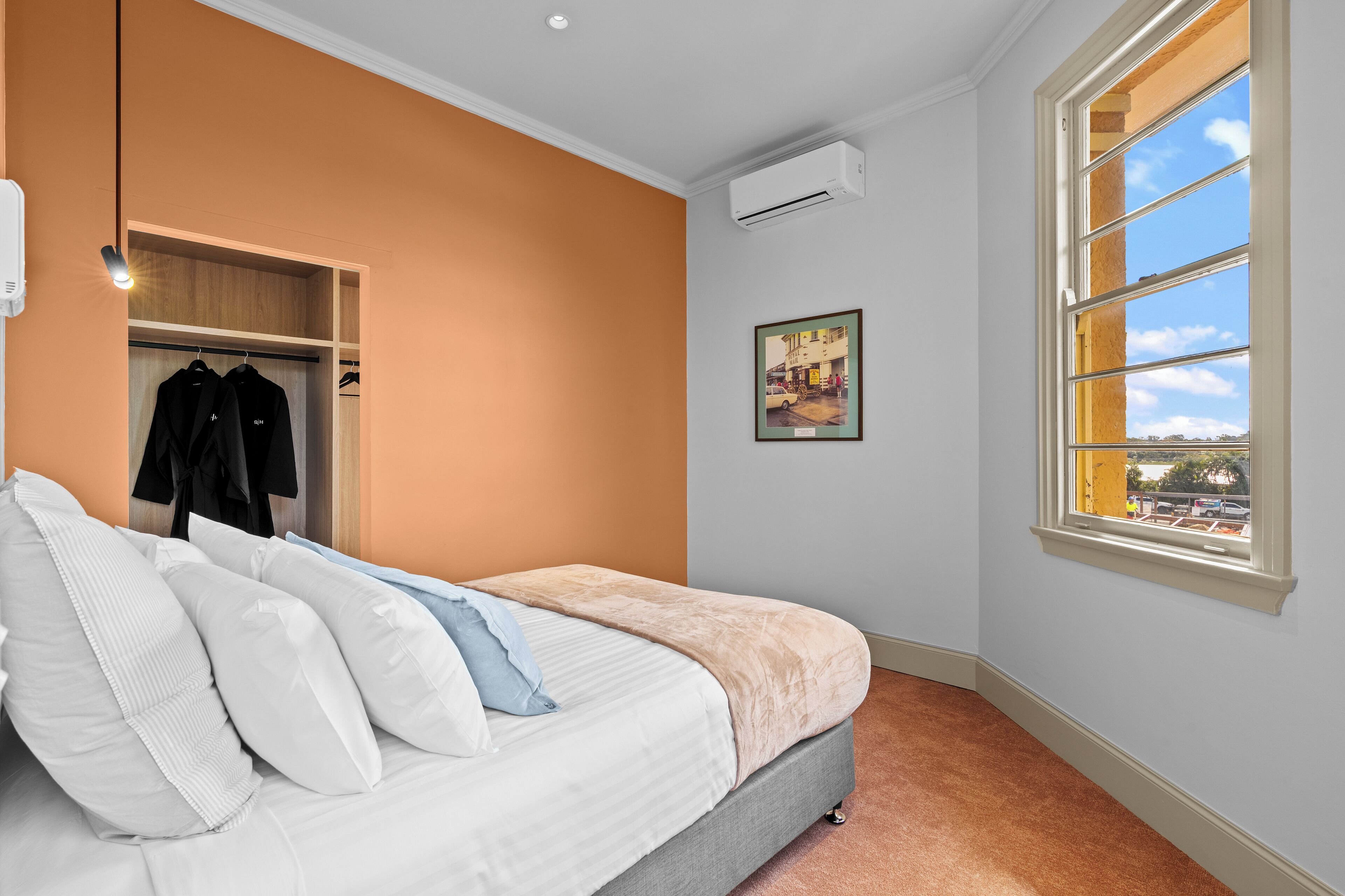 A bedroom with a bed, a closet, an air conditioning unit, a window with a view, and a framed picture on the wall.