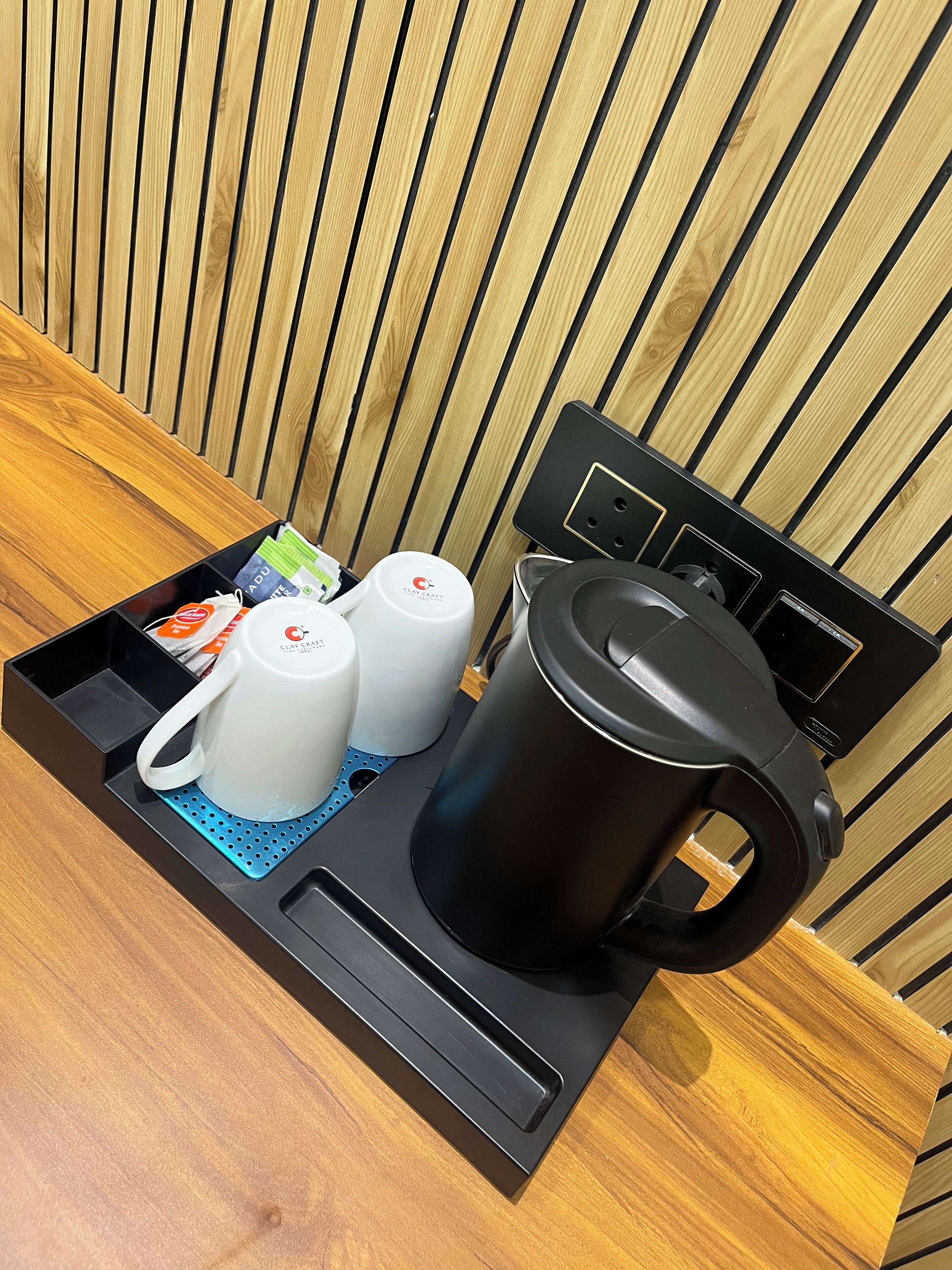 A coffee maker, two white cups, and a tray with tea bags and a box of tissues on a wooden table against a wooden wall.