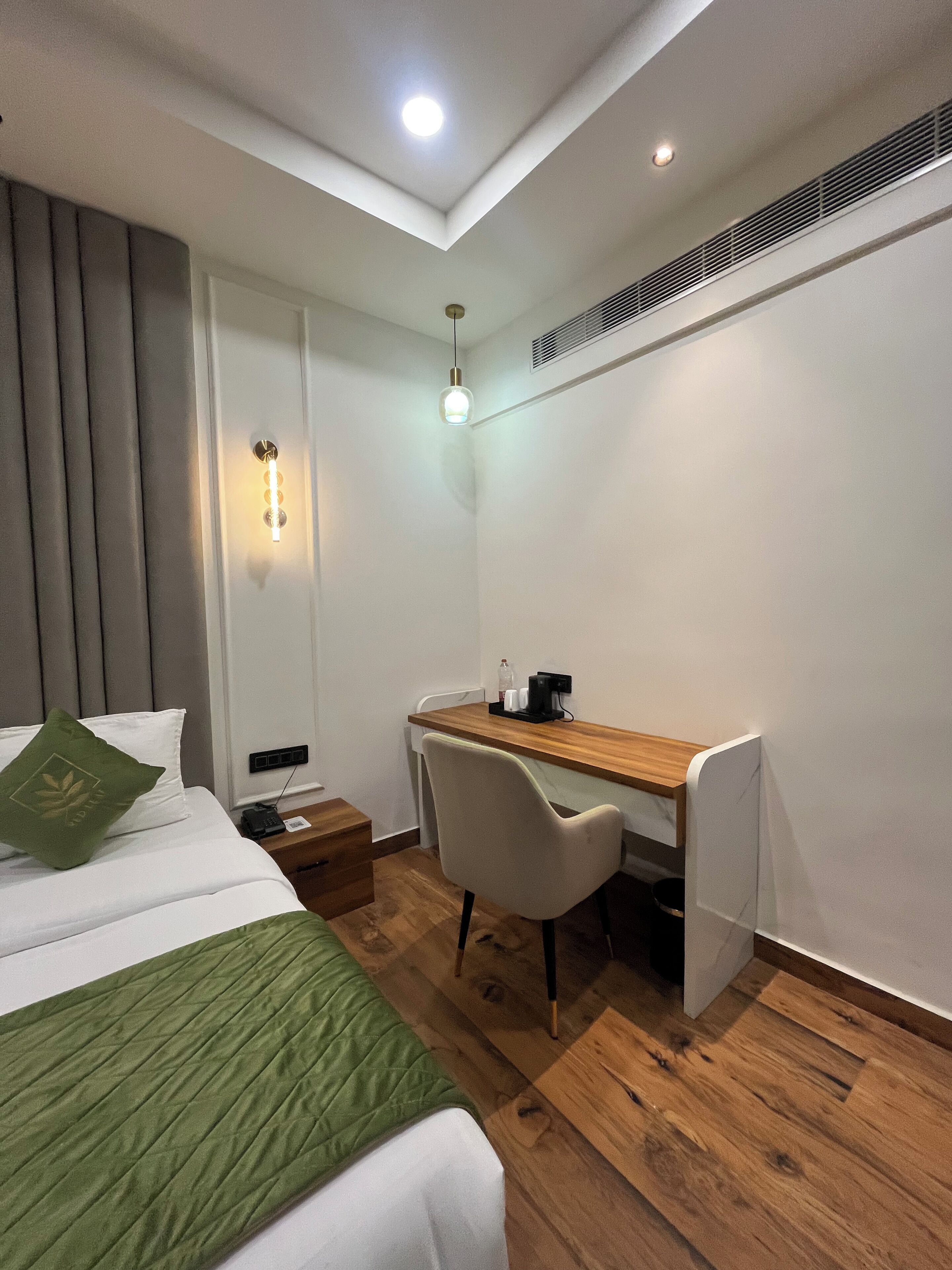A hotel room with a bed, a desk, a chair, a nightstand, and a wall-mounted light fixture.