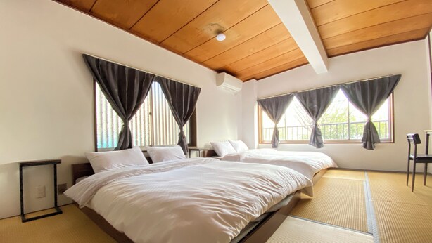 Habitación de hotel con una cama grande, dos ventanas con cortinas, techo de madera y esteras de tatami en el suelo.