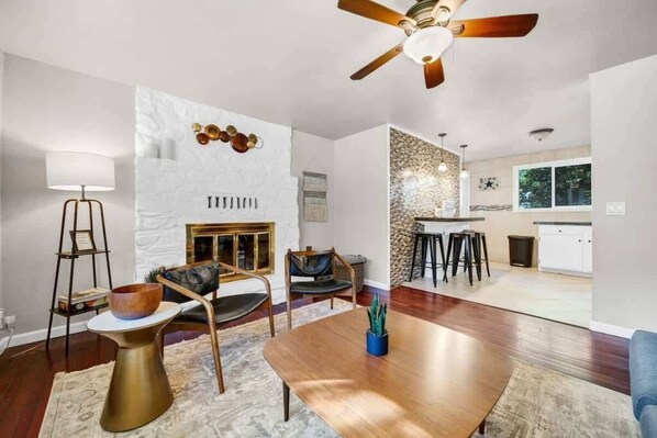 A modern living room with a wooden dining table, two chairs, a fireplace, and a kitchen in the background.