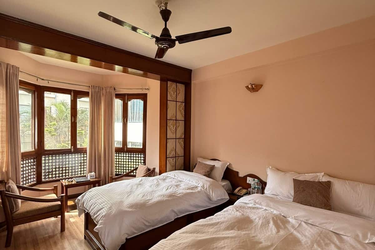 Habitación de hotel con dos camas, ventilador de techo, ventana con cortinas y vista a zonas verdes.