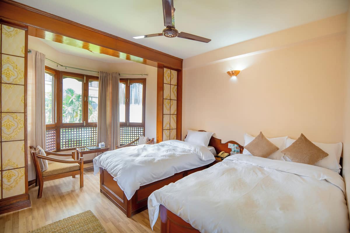 Habitación de hotel con dos camas, un ventilador de techo de madera, una silla y vistas a palmeras a través de las ventanas.