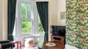Una sala de estar con un sofá verde, una mesita, un televisor sobre un mueble de madera y una ventana con cortinas.