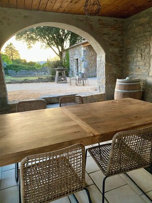 Un espace de restauration en plein air avec un mur en pierre, une table en bois, des chaises en osier et une vue sur un bâtiment en pierre et des arbres.