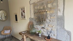 Une table en bois rustique, ornée d’un vase vert, d’une petite plante et d’un chandelier. Une chaise en osier avec un coussin à motifs se trouve à gauche. Un mur de pierre, décoré d’objets suspendus et surmonté d’un miroir, constitue le fond.