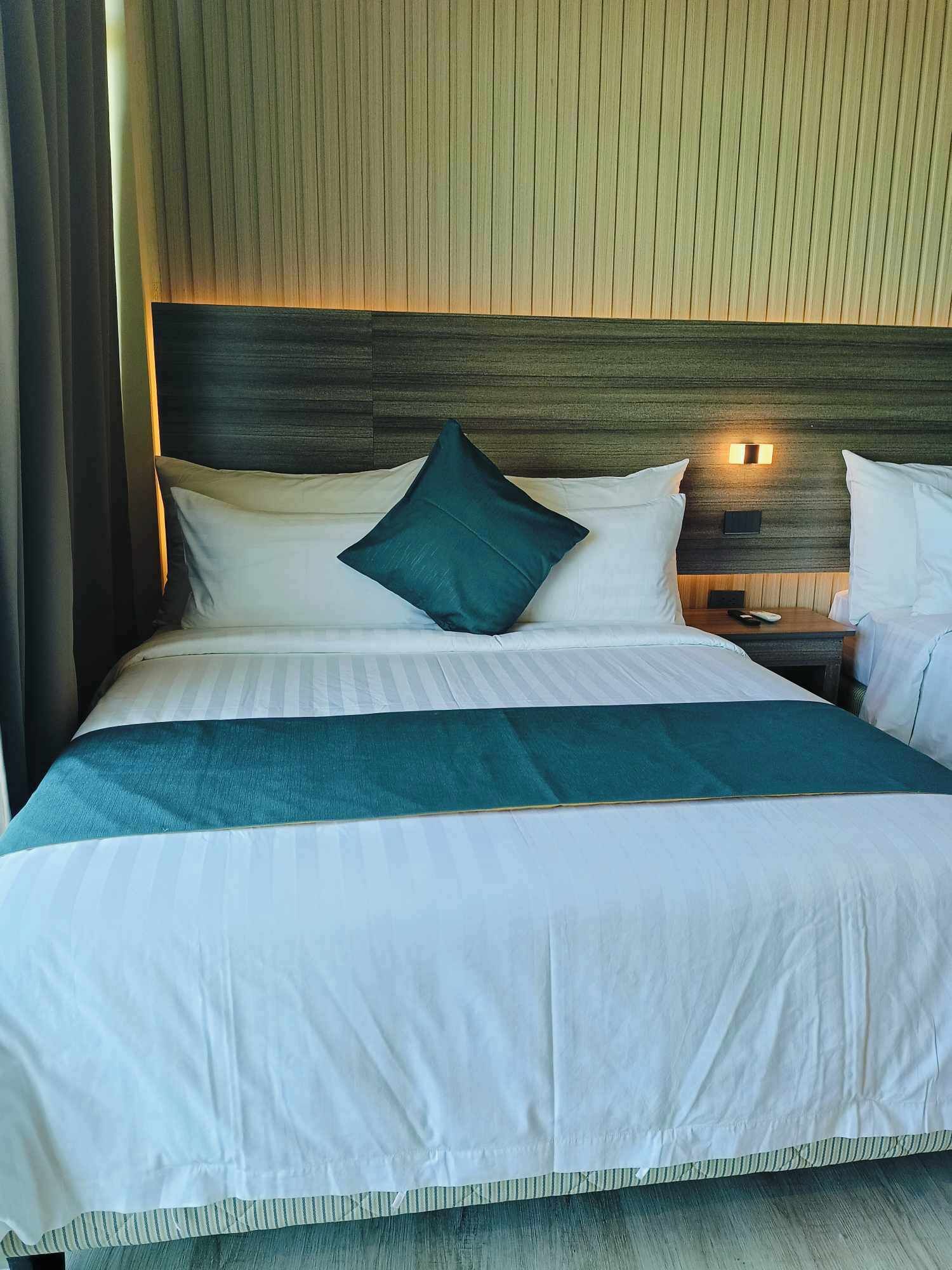 A neatly made bed with white linens and a teal throw blanket, a wooden headboard, and a bedside table with a lamp.