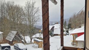 A wooden porch with two wooden chairs, a view of a snowy street with parked cars, and houses in the background.