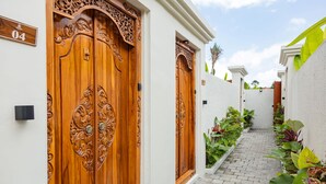 A narrow brick pathway leading to a wooden door with intricate carvings, a white wall with a number sign, and a small garden with various plants. - Jenna Villa by GenuineHost (Ubud)