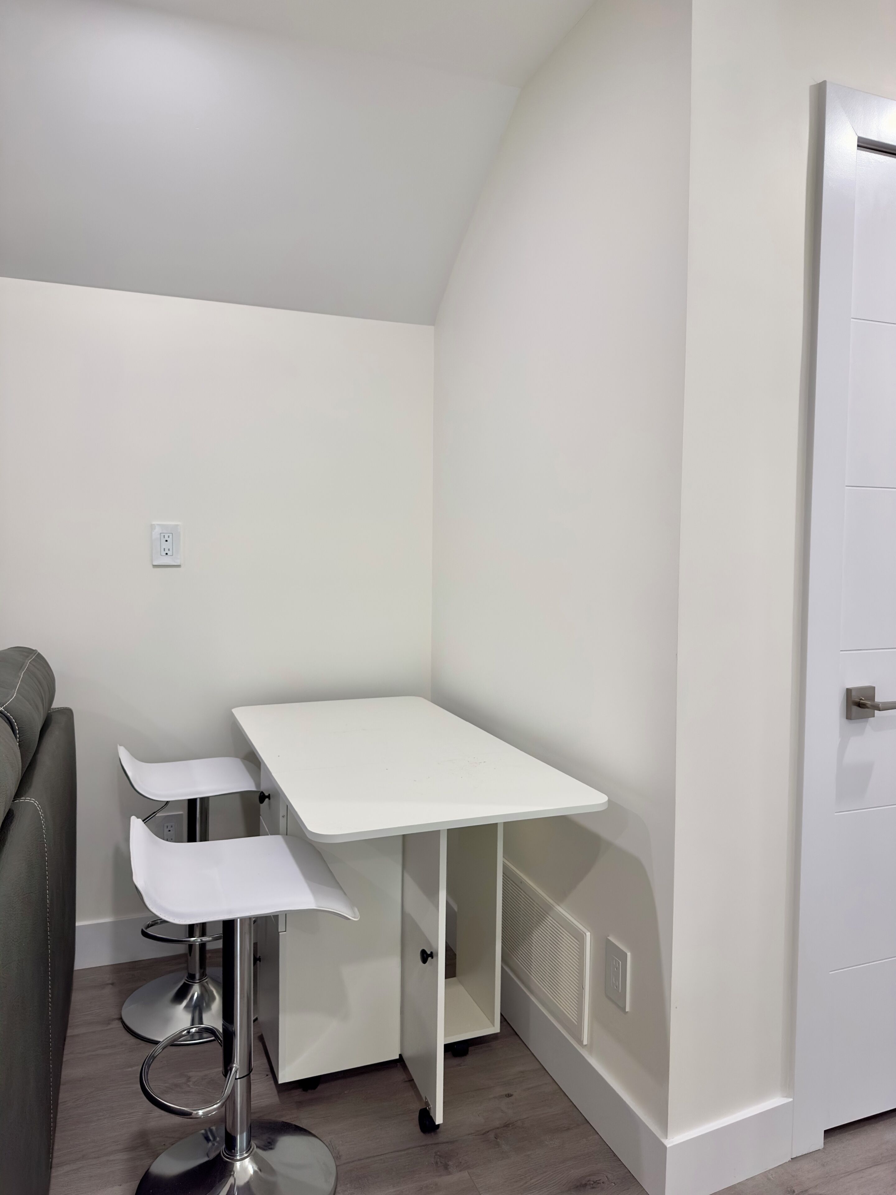 Una habitación en esquina con una mesa blanca y dos taburetes altos, un sofá gris y una puerta blanca.