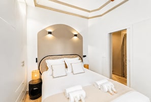 A modern bedroom with a large bed, a bedside table, a lamp, and a door leading to another room.