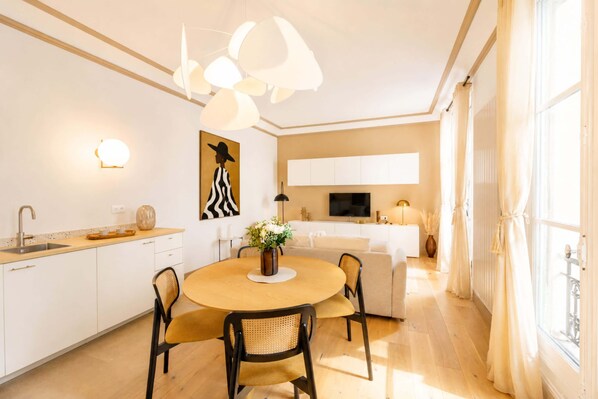 Une cuisine moderne avec des meubles blancs, un coin repas doté d’une table ronde et de chaises, et une grande fenêtre agrémentée de rideaux.