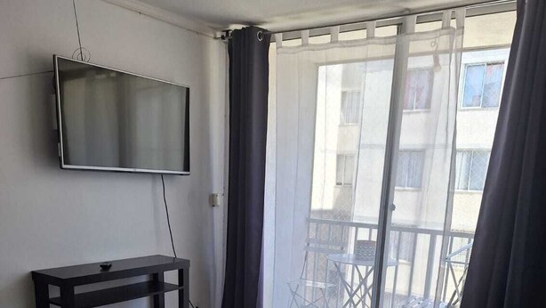 A living room with a black sofa, a TV mounted on the wall, a small table, and a sliding glass door leading to an outdoor area.