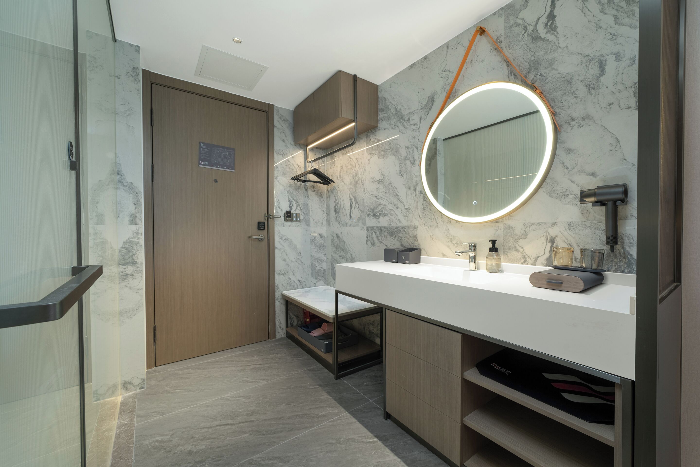 A modern bathroom with a marble backsplash, a round mirror with a light, and built-in cabinets.