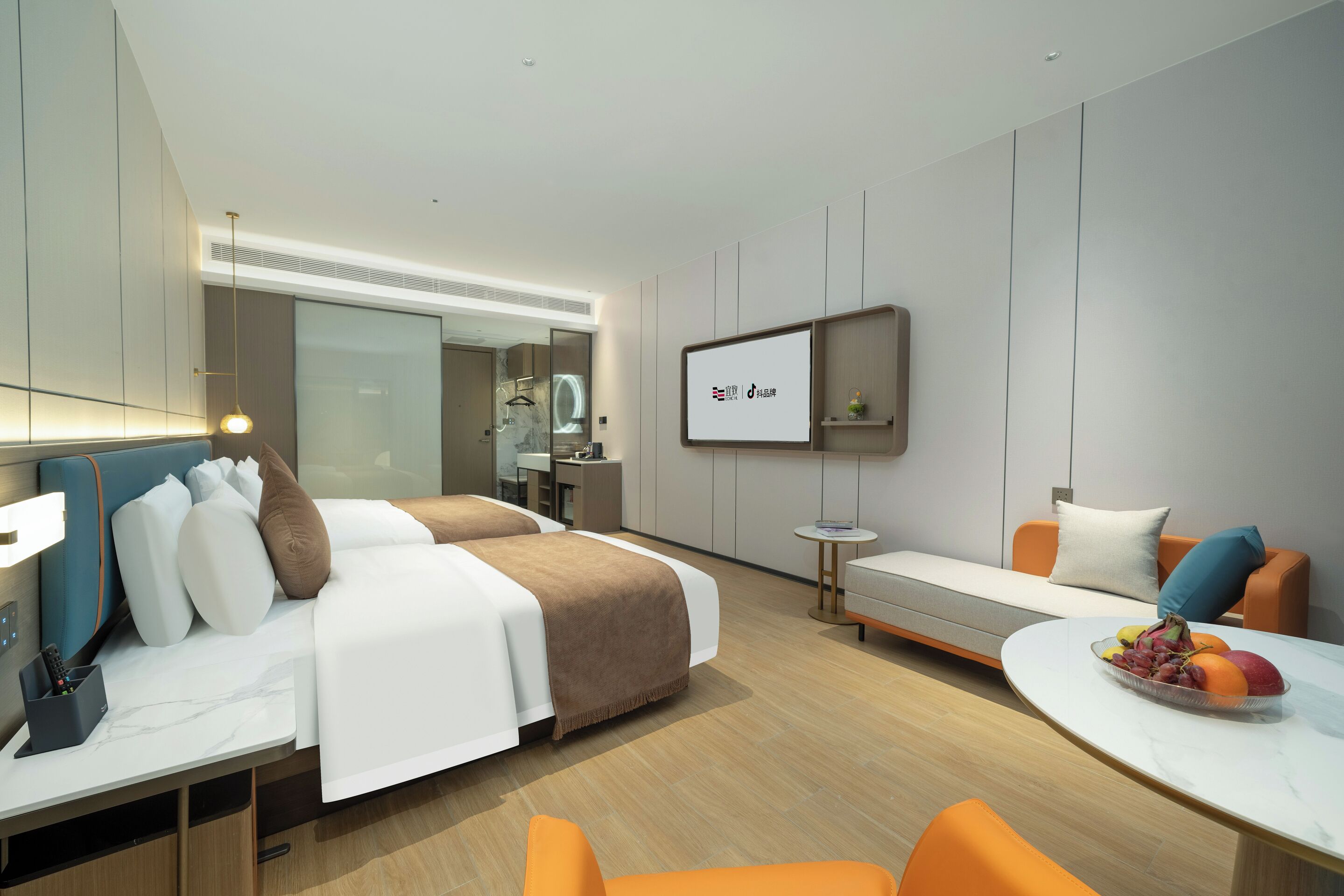 A modern hotel room with a bed, a desk, a seating area, and a TV screen displaying a logo.
