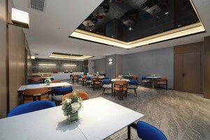 Un'area pranzo moderna con diversi tavoli e sedie, un composizione floreale al centro e un soffitto alto con illuminazione a incasso.