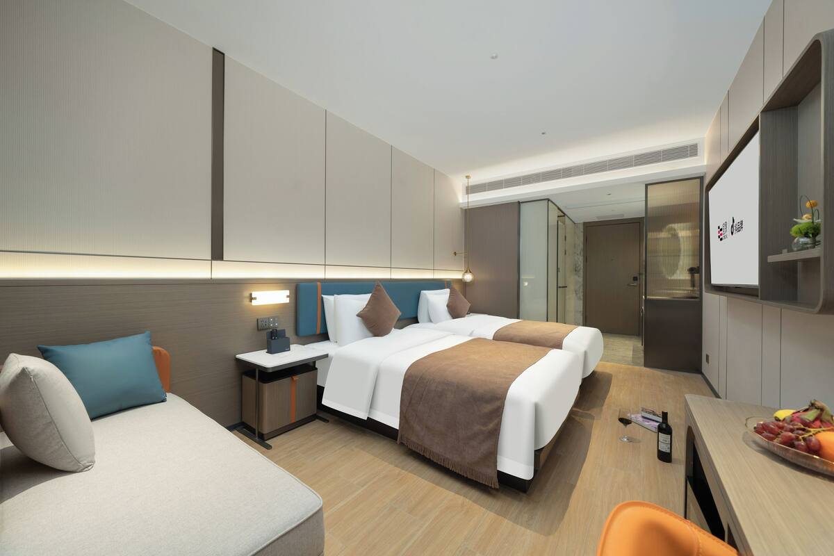 A modern hotel room with two beds, a desk, a chair, a minibar, and a small dining area with fruit.