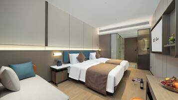 A modern hotel room with two beds, a desk, a chair, a minibar, and a small dining area with fruit.