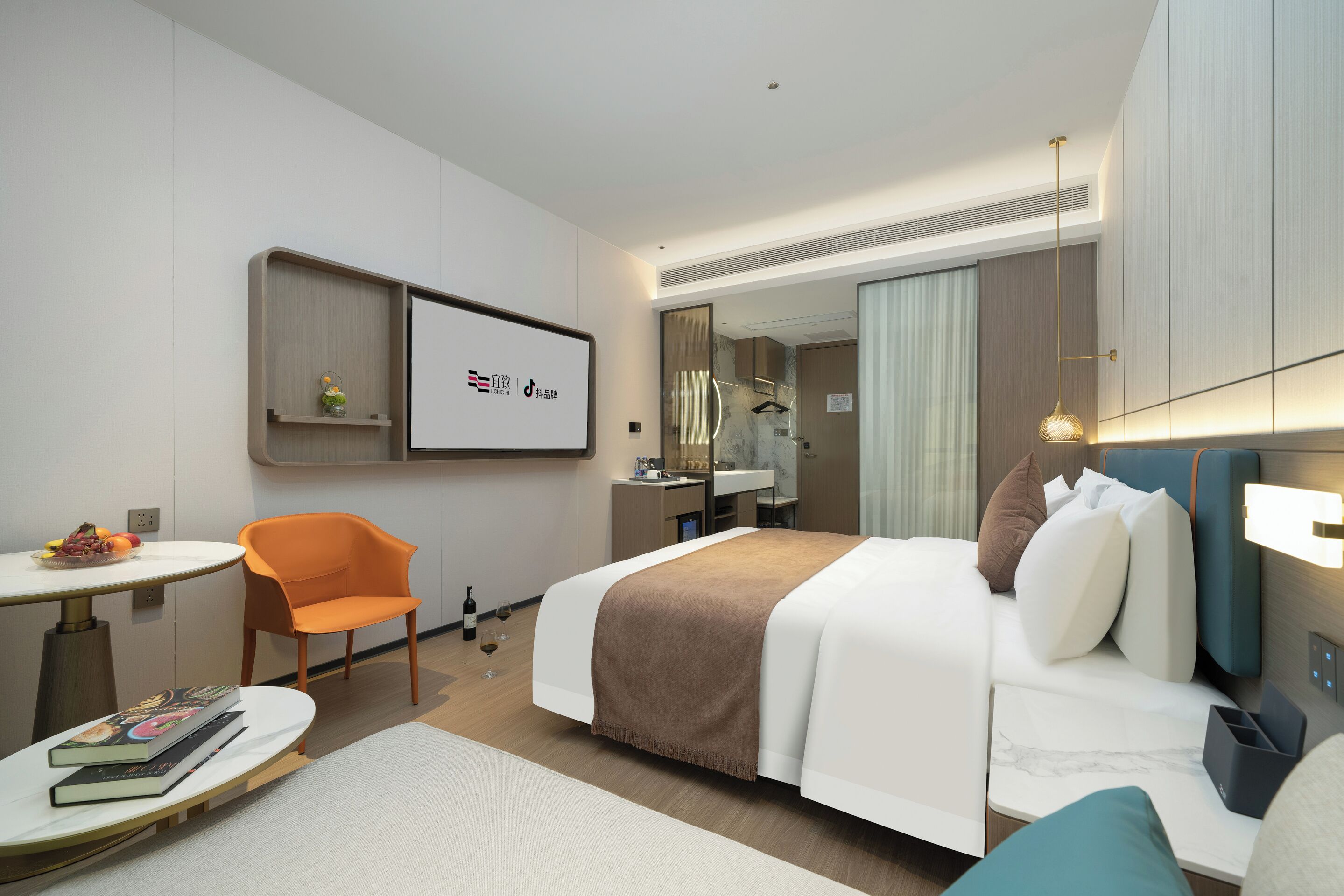 A modern hotel room with a bed, a desk, a chair, a small table with books, and a mirror.