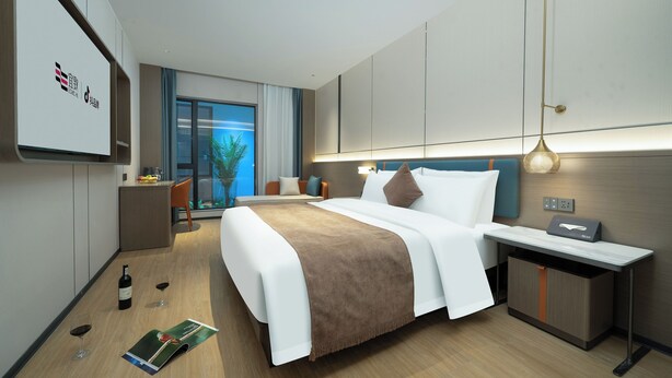 A modern hotel room with a large bed, bedside table, and a desk with a chair. There is a window with a view of palm trees and a balcony. - Yizhi Hotel Shamian Pearl River Night Cruise-Canton Fair free shuttle bus included free Breakfast&Gym&Laundry&free coffee allday (Guangzhou)