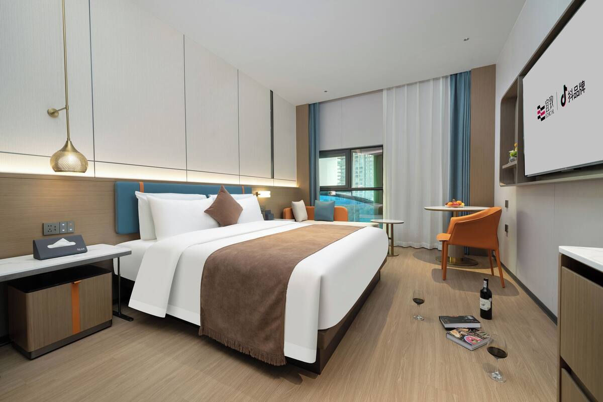 A modern hotel room with a large bed, bedside table, and a desk with a chair. There is a window with curtains, a small table with a chair, and a TV mounted on the wall.